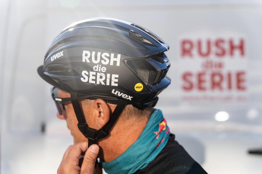 Cyclist with a black "RUSH the SERIES" helmet in profile, fastening the chin strap.